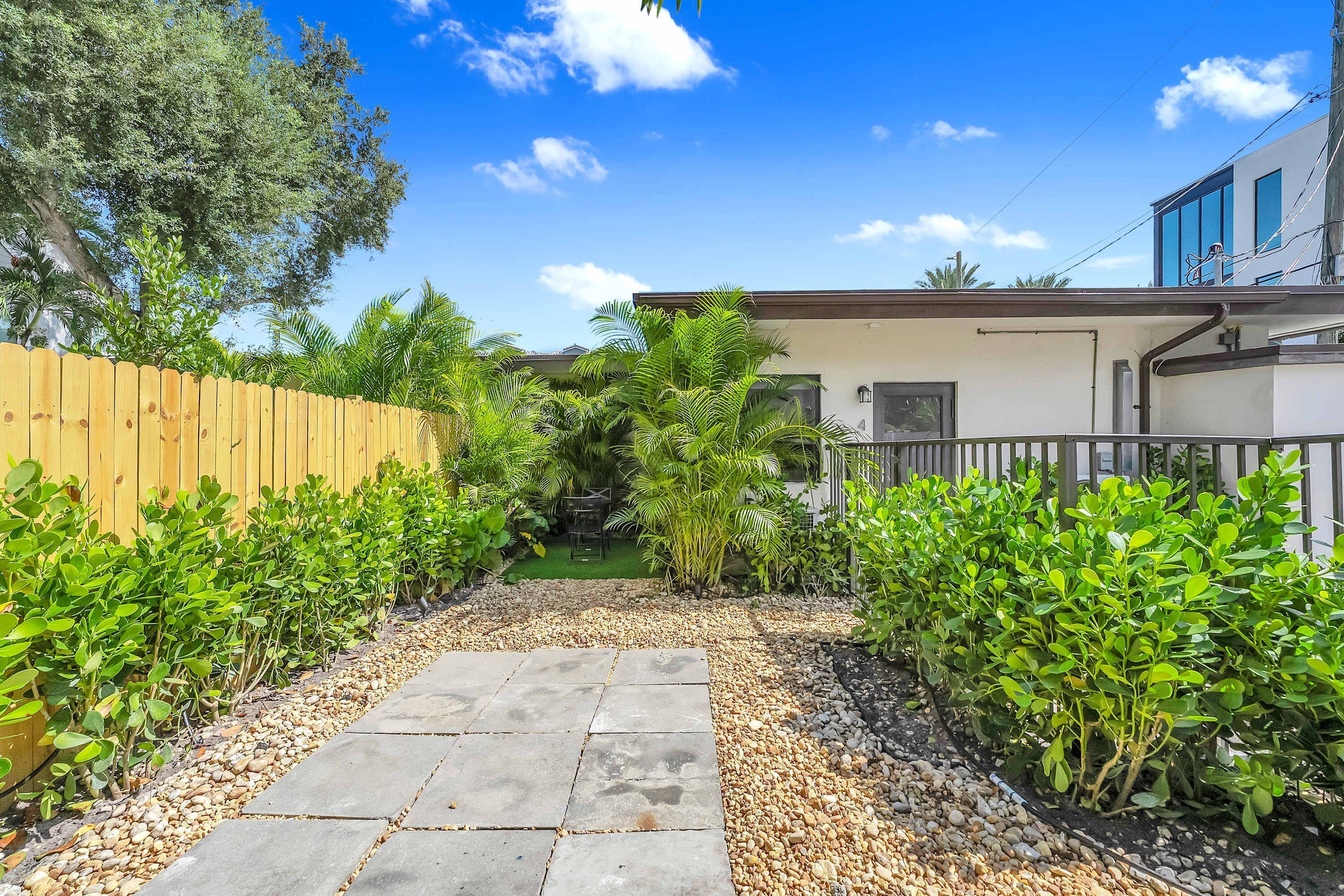 A pathway leading to a house with a wooden fence on one side and a metal fence on the other, surrounded by lush greenery and a clear blue sky.