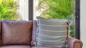 A modern living room with a brown leather sofa, a round glass coffee table, and a view of lush green palm trees through large windows. - Chic Modern Studio With Queen Bed (Fort Lauderdale)