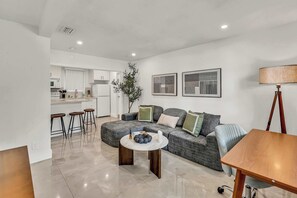 A modern living room with a grey sectional sofa, a round coffee table, and a wooden side table. There is a kitchen in the background with white cabinets and a dining area with bar stools. - Bright 1BR Getaway Victoria Park Las Olas Nearby (Fort Lauderdale)