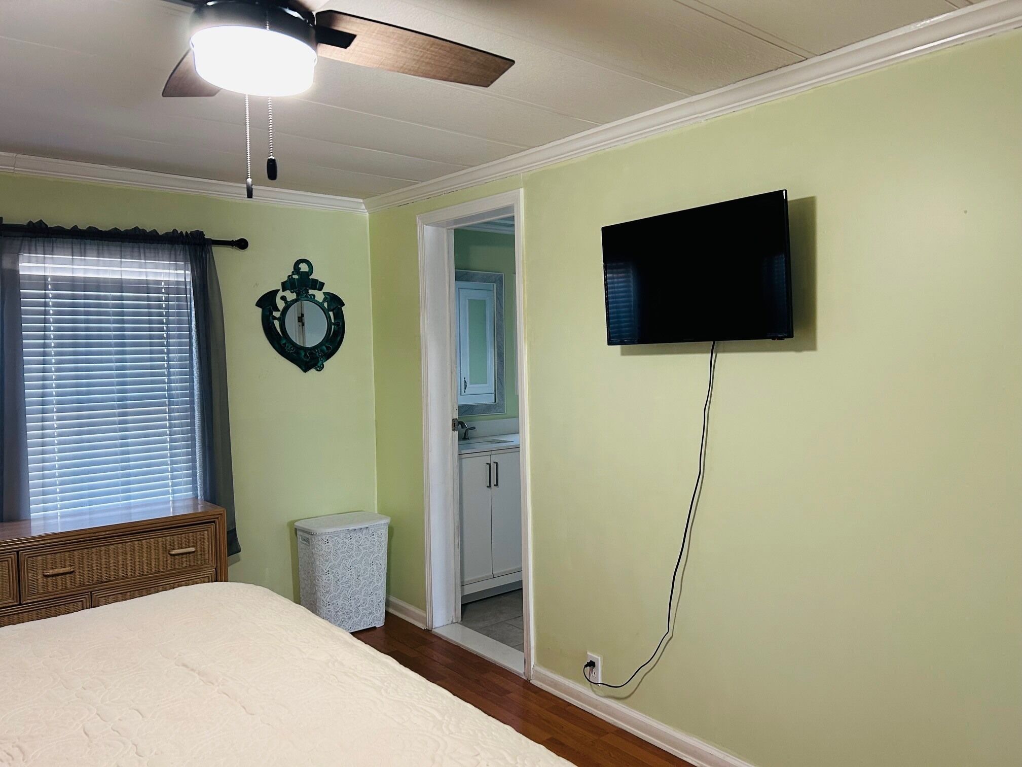 Quarto com cama, ventoinha no teto, TV fixada na parede, janela com cortinas e porta para um banheiro.