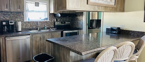 A kitchen with wooden cabinets, a granite countertop, a stainless steel dishwasher, a trash can, and wicker bar stools.