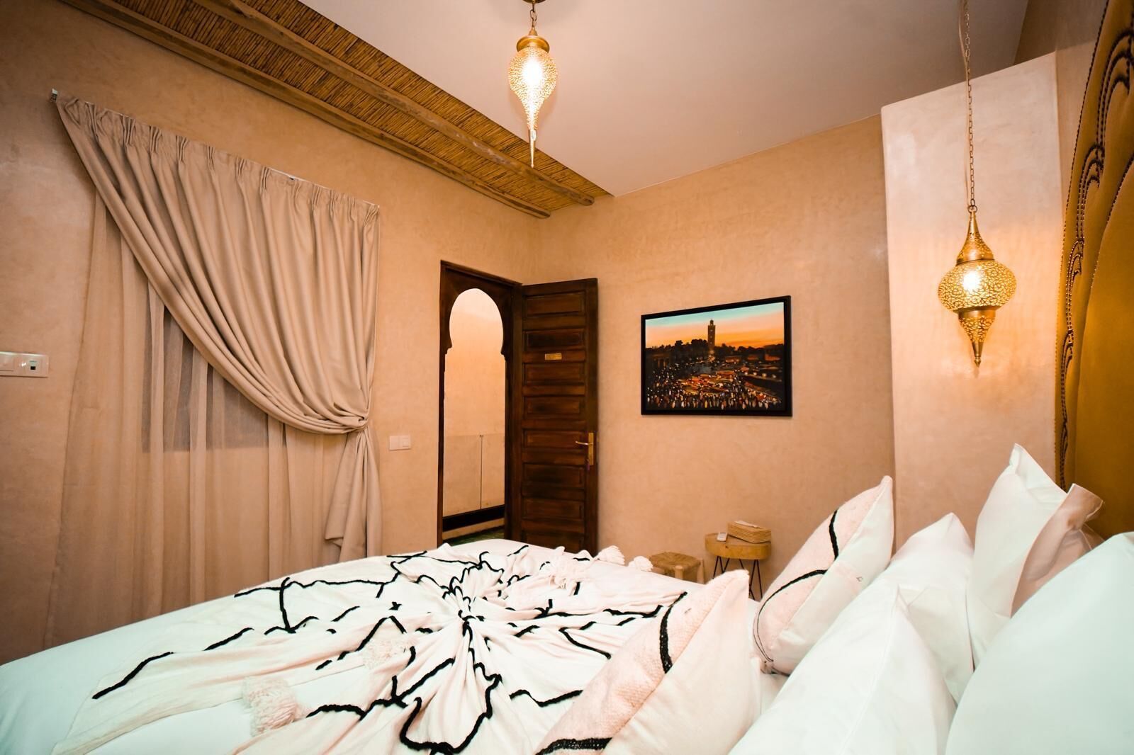A hotel room with a bed, a wooden door, a cityscape picture on the wall, and two hanging lamps.