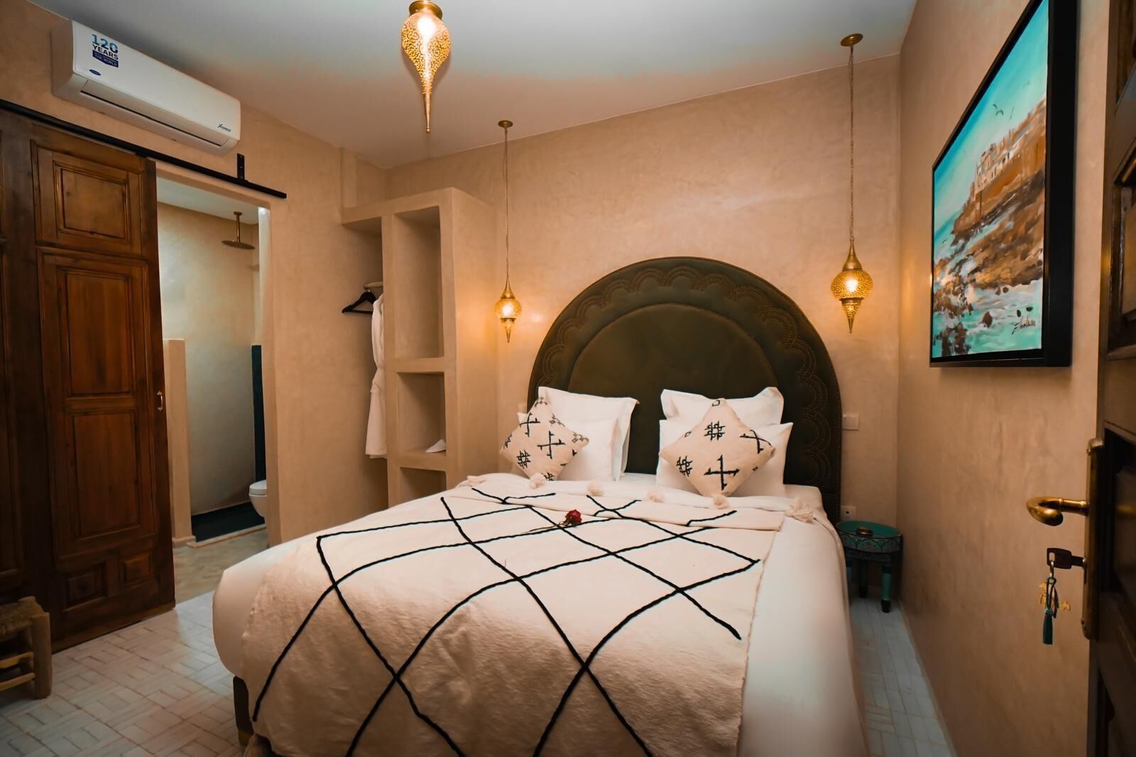 A bedroom with a large bed, a wooden wardrobe, a built-in shelf, a wall-mounted air conditioner, and a painting.