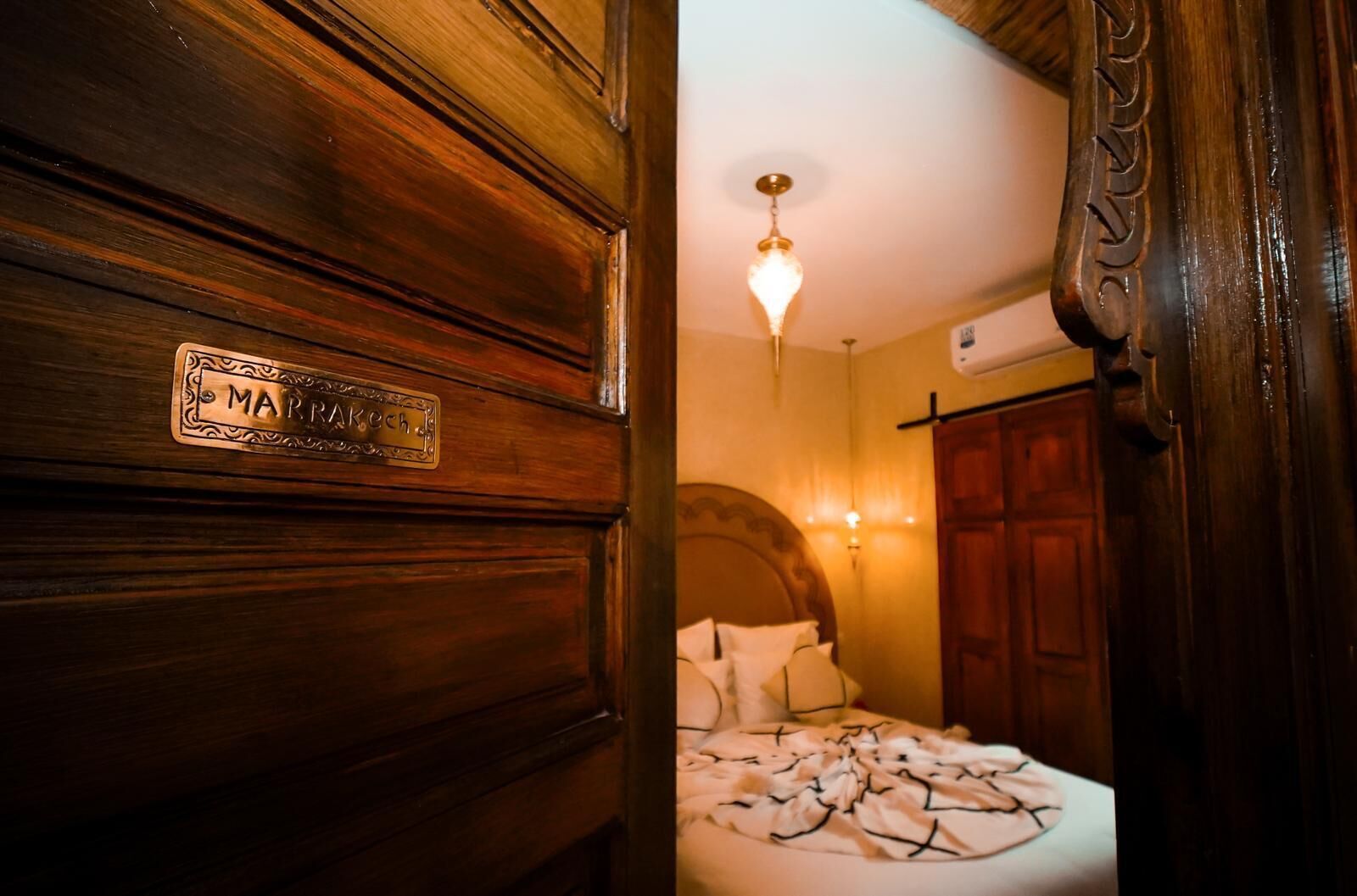A wooden door with a nameplate reading "MARRAKECH" and a bed with a white bedspread and pillows in a warmly lit room.