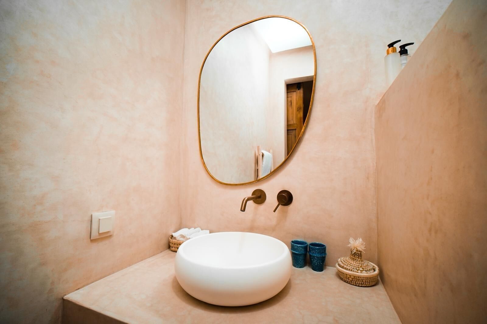 A bathroom with a round white sink, a gold-framed mirror, and a wooden door in the background.