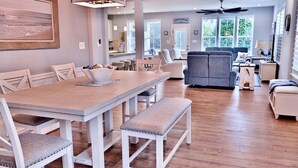 A spacious dining area with a wooden table and white chairs, a gray sofa, and a ceiling fan in a bright, well-lit room with large windows. - Bayside Resort: Great Location Walk to The Pool: (Selbyville)