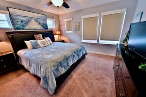 Un dormitorio con cama, televisor, mesita de noche, lámpara, ventana con persianas y ventilador de techo.