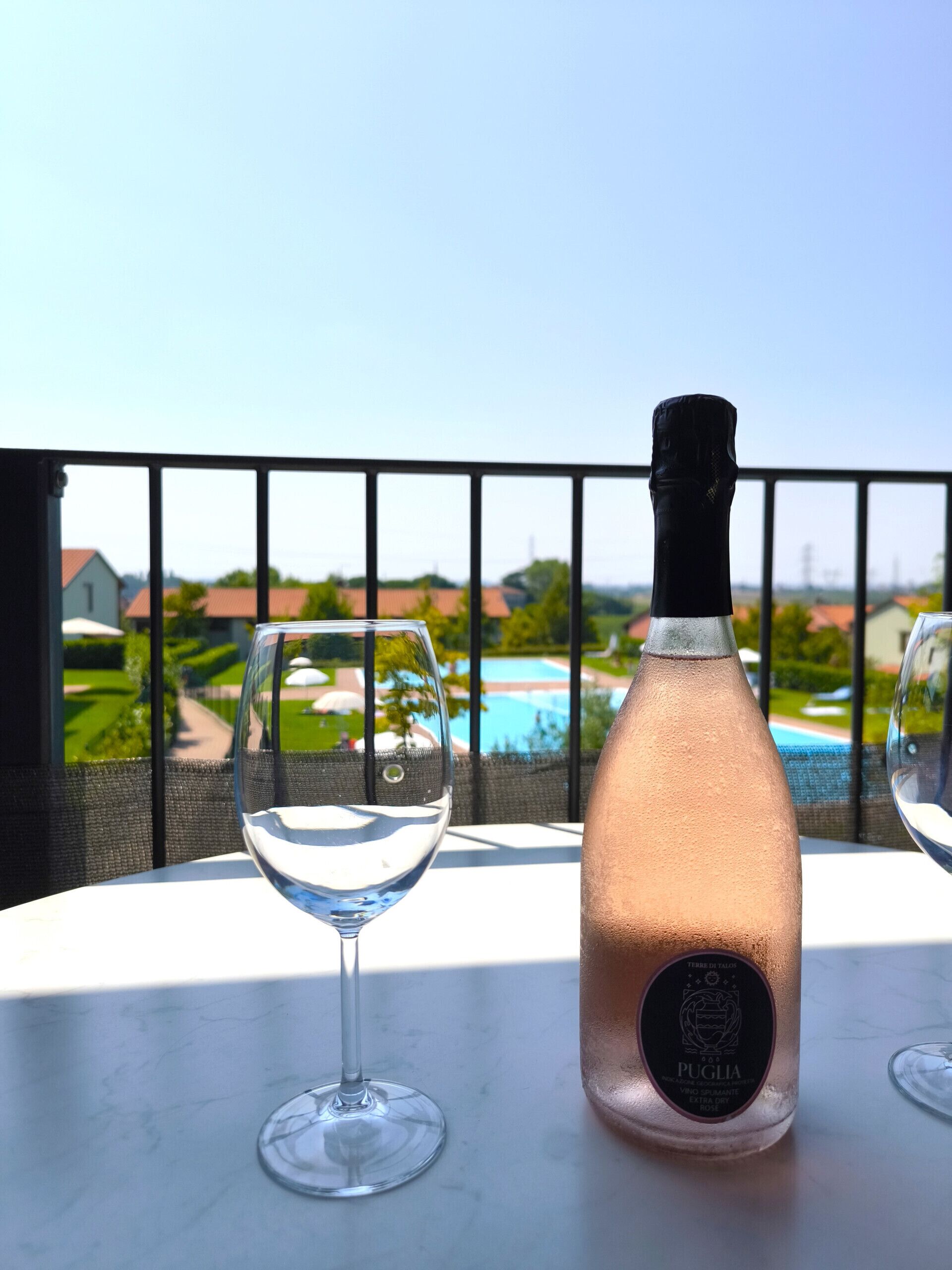 A bottle of Puglia wine and two empty wine glasses on a table with a view of a swimming pool and houses in the background.