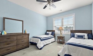 A bedroom with two beds, a dresser, a ceiling fan, a window with blinds, and a lamp on a nightstand.