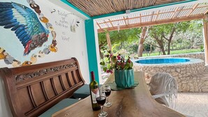 A wooden dining table with a bench, a bottle of wine, and glasses. There is a mural on the wall and a view of a garden and pool outside. - Private cabin with Jacuzzi, just one minute from the beach. (Bolívar)