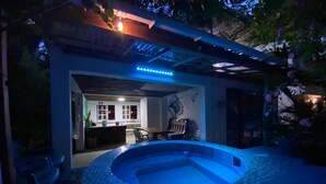 Una piscina circular con mosaicos azules, rodeada por un porche cubierto con asientos y una mesa.