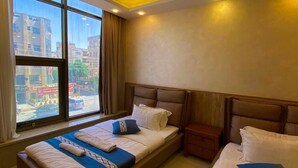 A hotel room with two beds, a nightstand, a desk, and a large window with a city view. - almansi Hotels (Amman)
