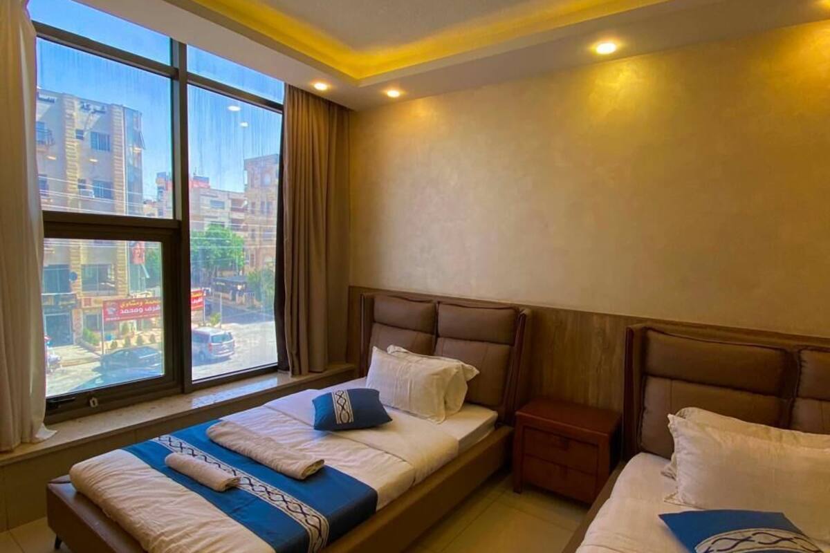 A hotel room with two beds, a nightstand, a desk, and a large window with a city view.