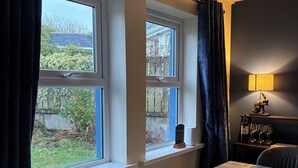 Habitación con vistas al jardín a través de amplios ventanales, una mesa con una silla, un radiador y una lámpara en un estante.