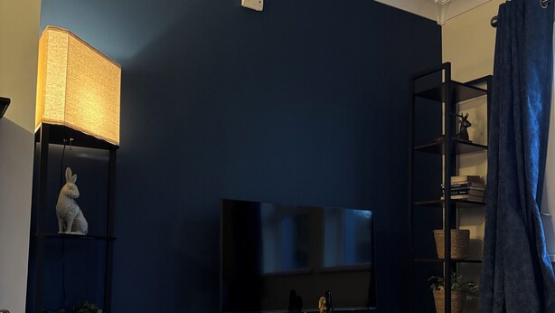 Una sala con paredes azul oscuro, piso de madera, televisor sobre un mueble, lámpara, estante con objetos decorativos y cortina azul.