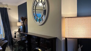 Una habitación con chimenea, un reloj y una mesa con lámpara y objetos decorativos.
