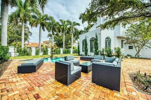 A patio with wicker furniture and a swimming pool, surrounded by palm trees and a white building. - Beautiful Historic Ringling house! (Sarasota)