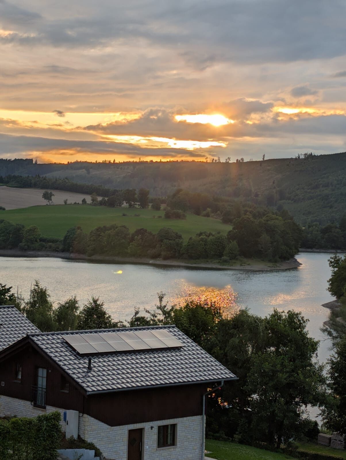 A house with a sloped roof and solar panels, overlooking a river and hilly landscape during sunset.