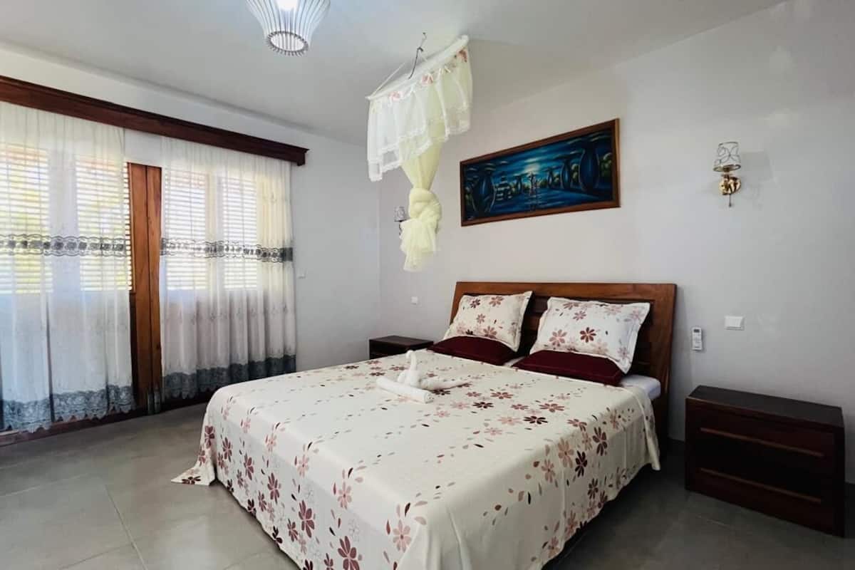 A bedroom with a large bed, two bedside tables, a nightstand, a window with curtains, and a wall-mounted lamp.