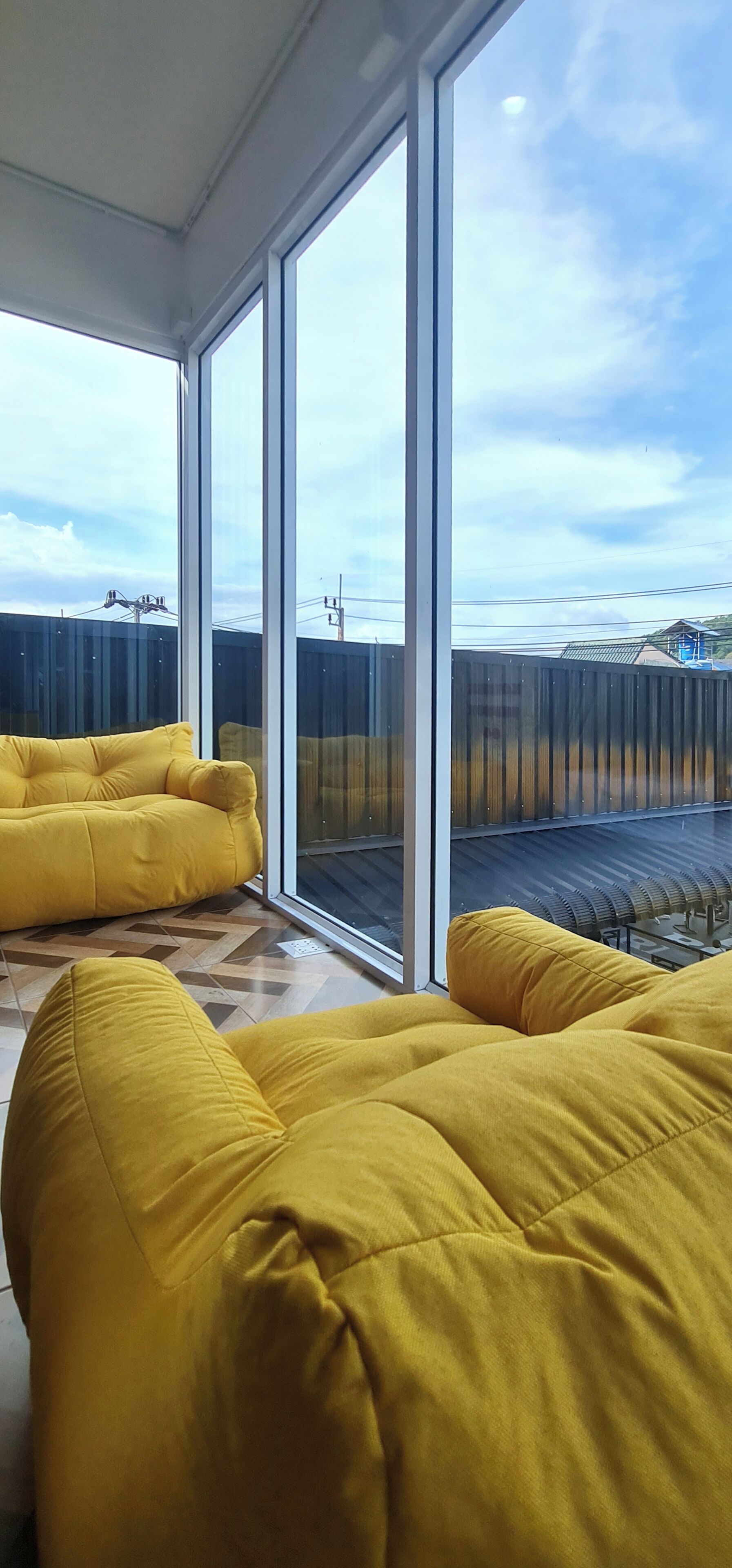 Una habitación con ventanas amplias, sofás amarillos y vista a un área cercada con edificios.