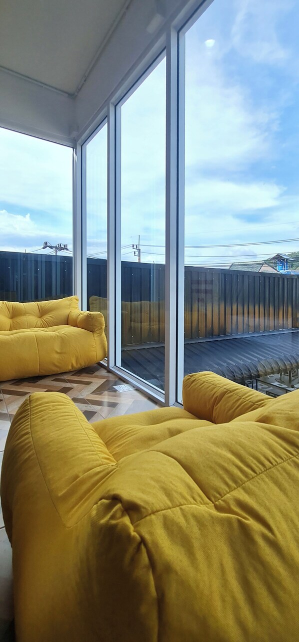 A room with large windows, yellow sofas, and a view of a fenced area with buildings. - Sleep On The Clouds (Karon)