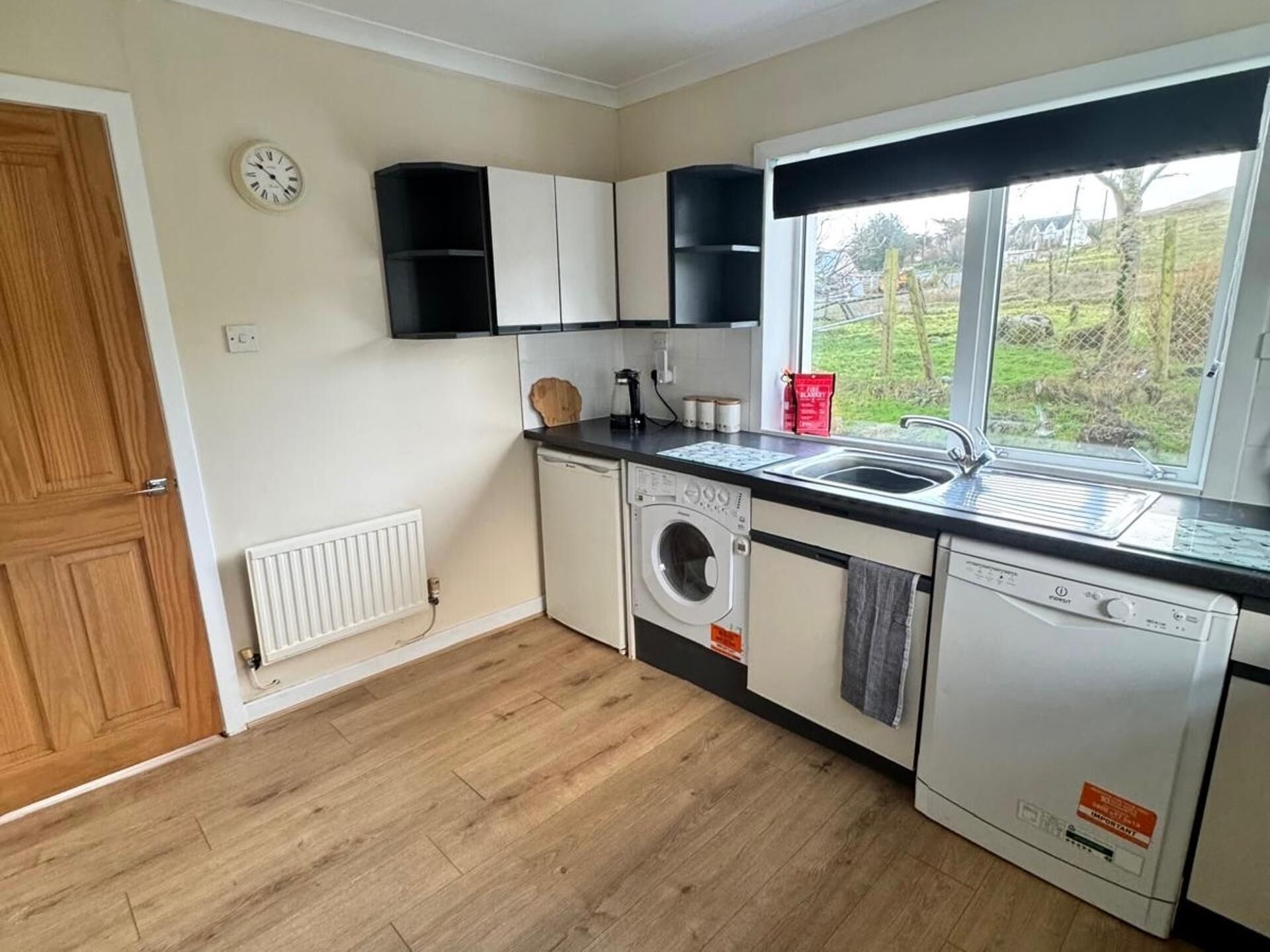 A compact kitchen with a washing machine, a dishwasher, and a window with a view of greenery.