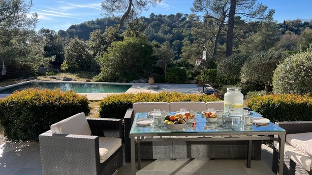 An outdoor dining area with a glass table and wicker chairs, overlooking a pool and lush greenery. - Modern architect-designed villa with pool - Immersion in nature & light (Roquefort-les-Pins)