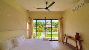 Un dormitorio con cama, ventilador de techo, ventana con cortinas, vista a una piscina y jardín, y una mesita con un florero.