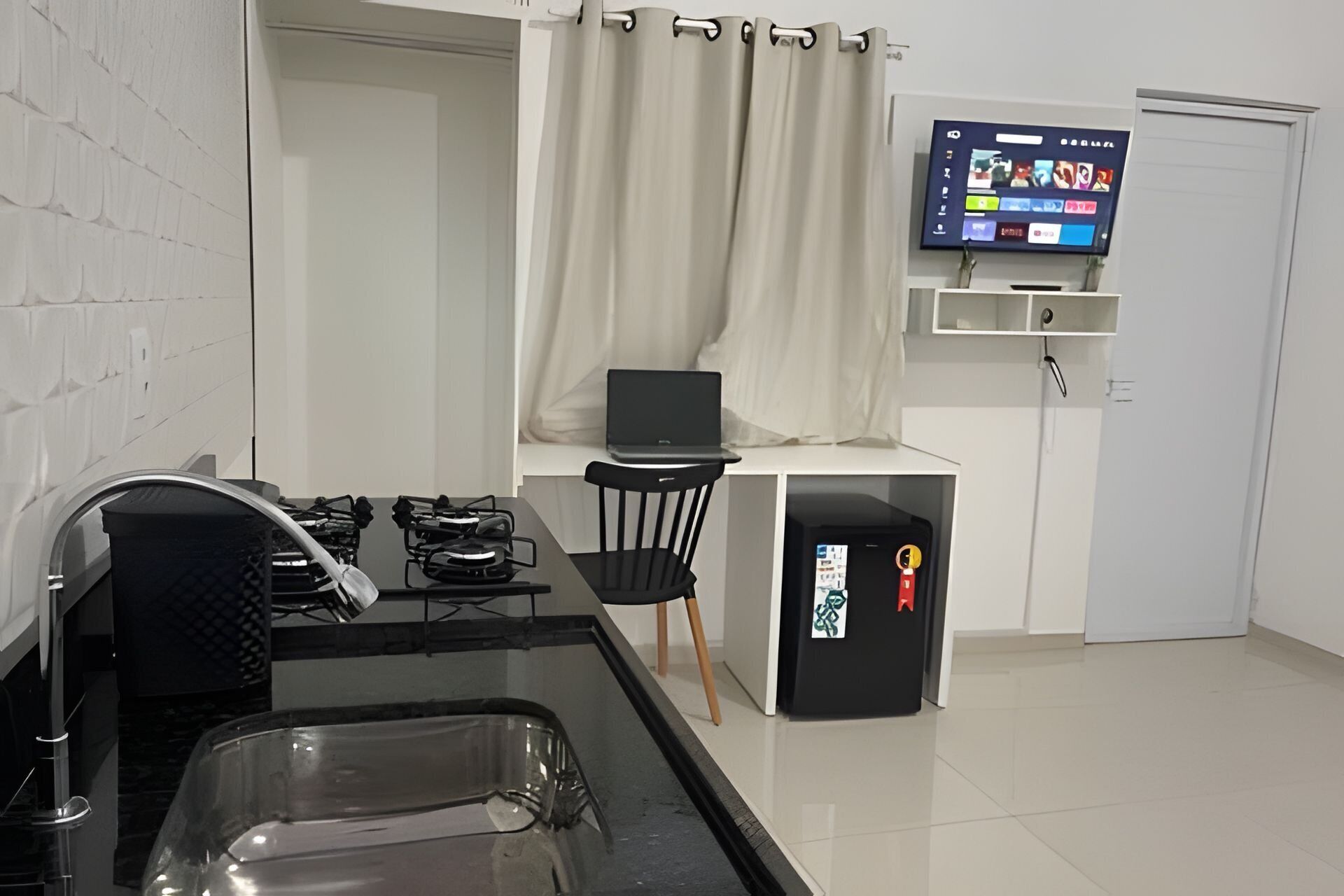 A modern kitchen with a gas stove, a sink, and a black countertop. There is a laptop on a small table, a TV mounted on the wall, and a black cabinet with a red sticker.