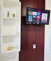 Televisão fixada na parede exibindo vários canais e uma prateleira com objetos decorativos.