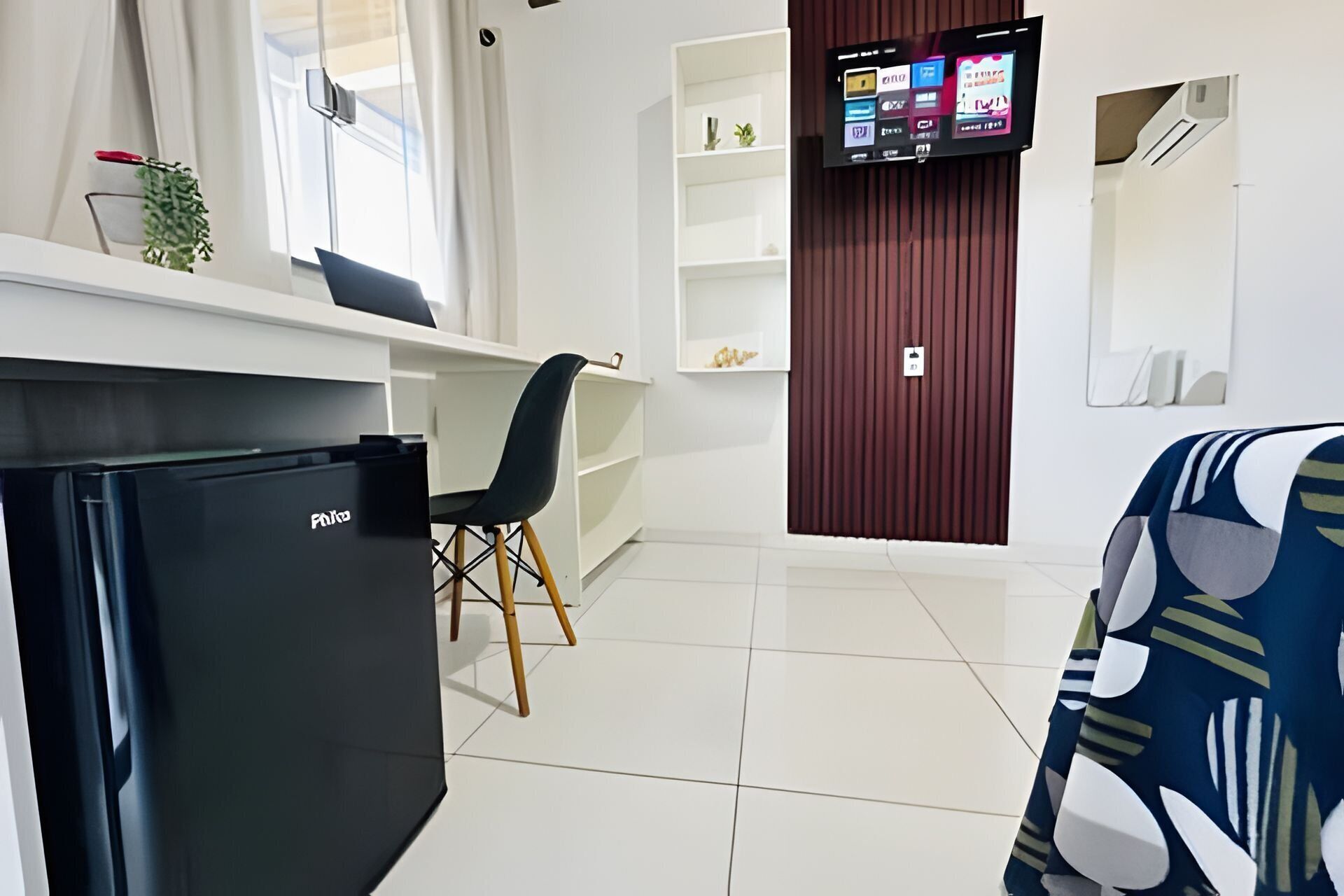Quarto de hotel moderno com um frigorífico preto, uma escrivaninha com cadeira, uma televisão na parede e um sofá estampado.