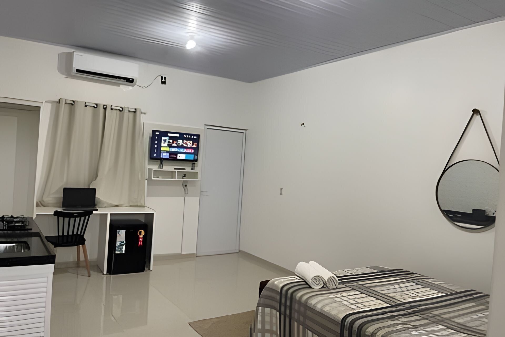 A hotel room with a bed, a desk with a laptop, a TV on a stand, and a small kitchenette.