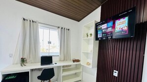 A room with a white desk, a black chair, a TV mounted on a wooden panel, and a window with curtains. - Apartamentos Vitória (Barreirinhas)