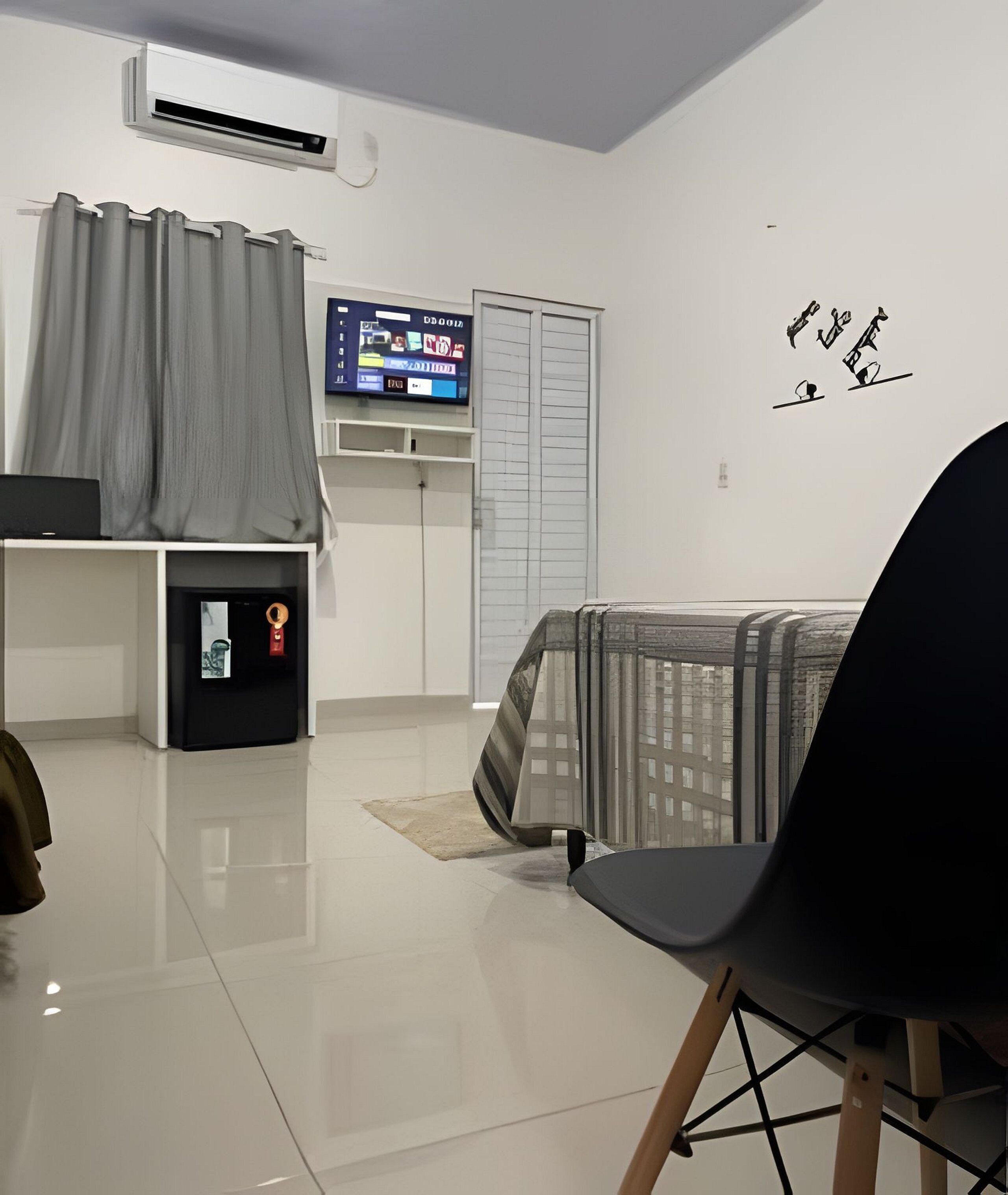 A modern living room with a flat-screen TV, a small refrigerator, a grey curtain, a bed with a striped blanket, and a wall with a decorative sign.