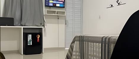 Una sala moderna con un televisor de pantalla plana, un refrigerador pequeño, una cortina gris, una cama con una manta a rayas y una pared con un letrero decorativo.