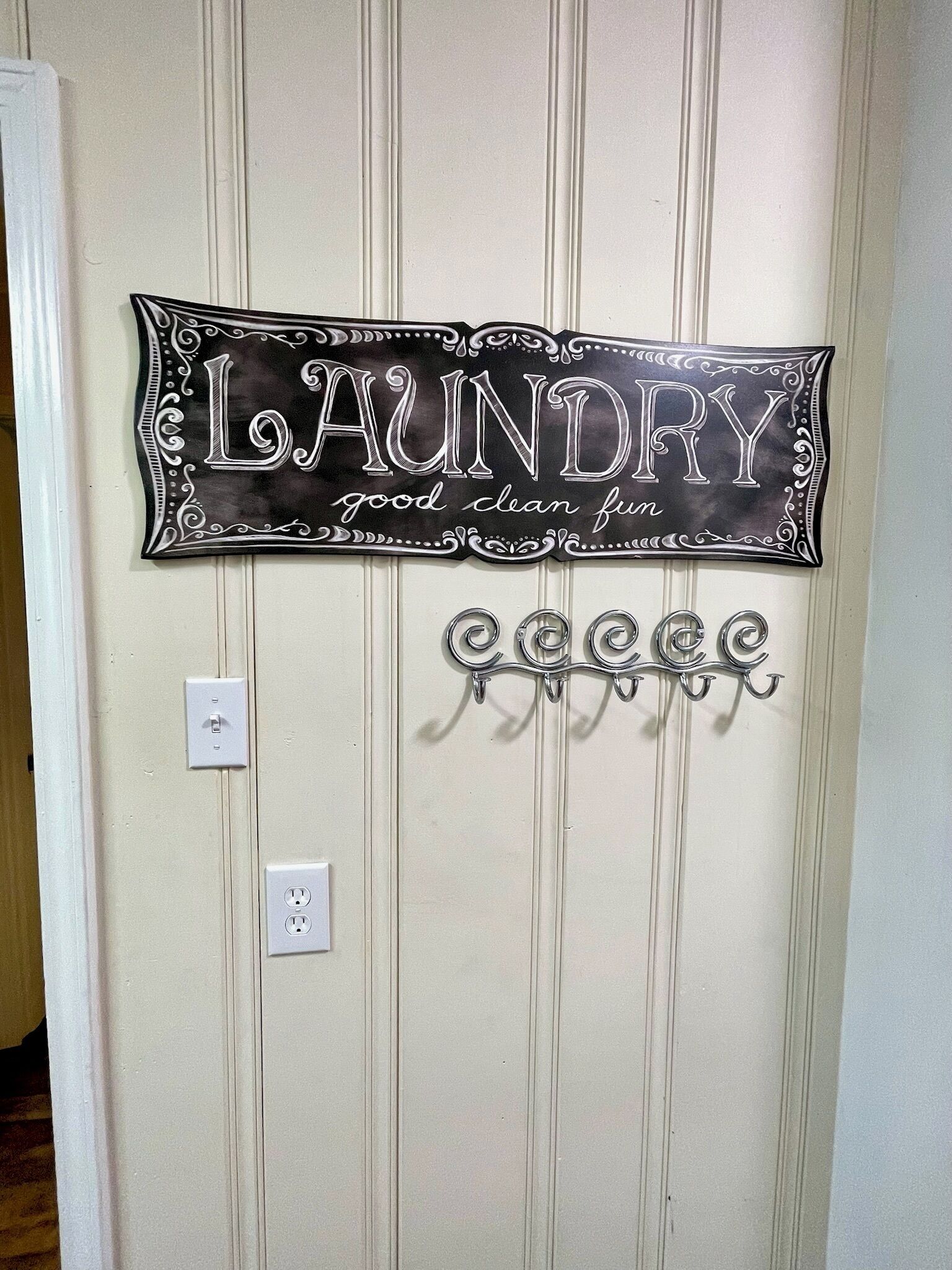 A wall-mounted sign with the word "LAUNDRY" and the phrase "good clean fun" in a decorative font. Below the sign, there are metal hooks for hanging items.