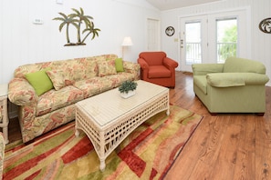 Un salon avec un canapé à motifs floraux, un fauteuil rouge, un fauteuil vert, une table basse en osier et un tapis à motifs.