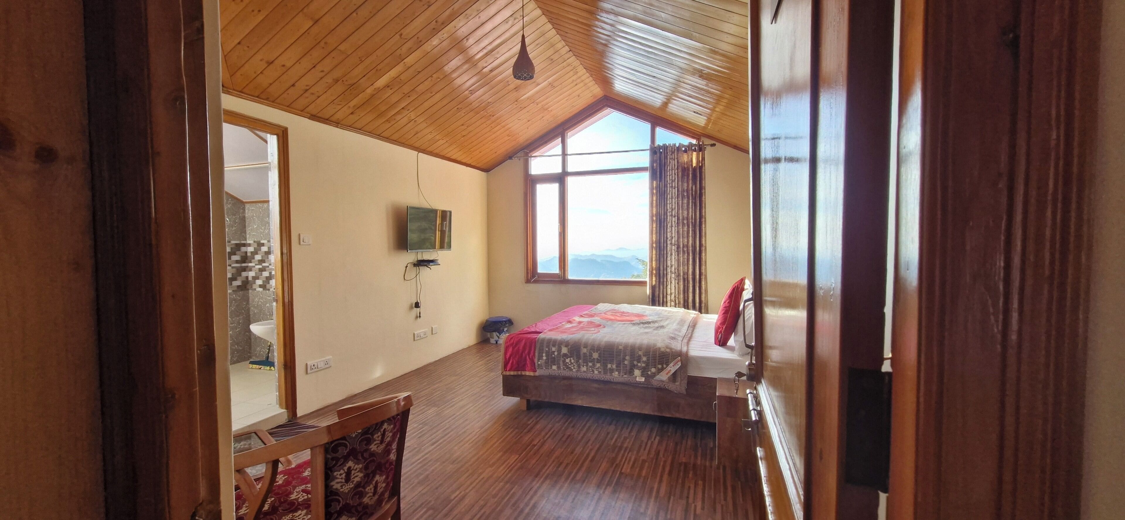 A bedroom with a wooden ceiling, a bed with a patterned bedspread, a bedside table, a mirror, and a window with a view.