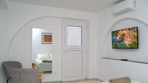 Een moderne hotelkamer met een bed, een stoel, een klein tafeltje met fruit, een televisie en een deur die naar een andere kamer leidt.