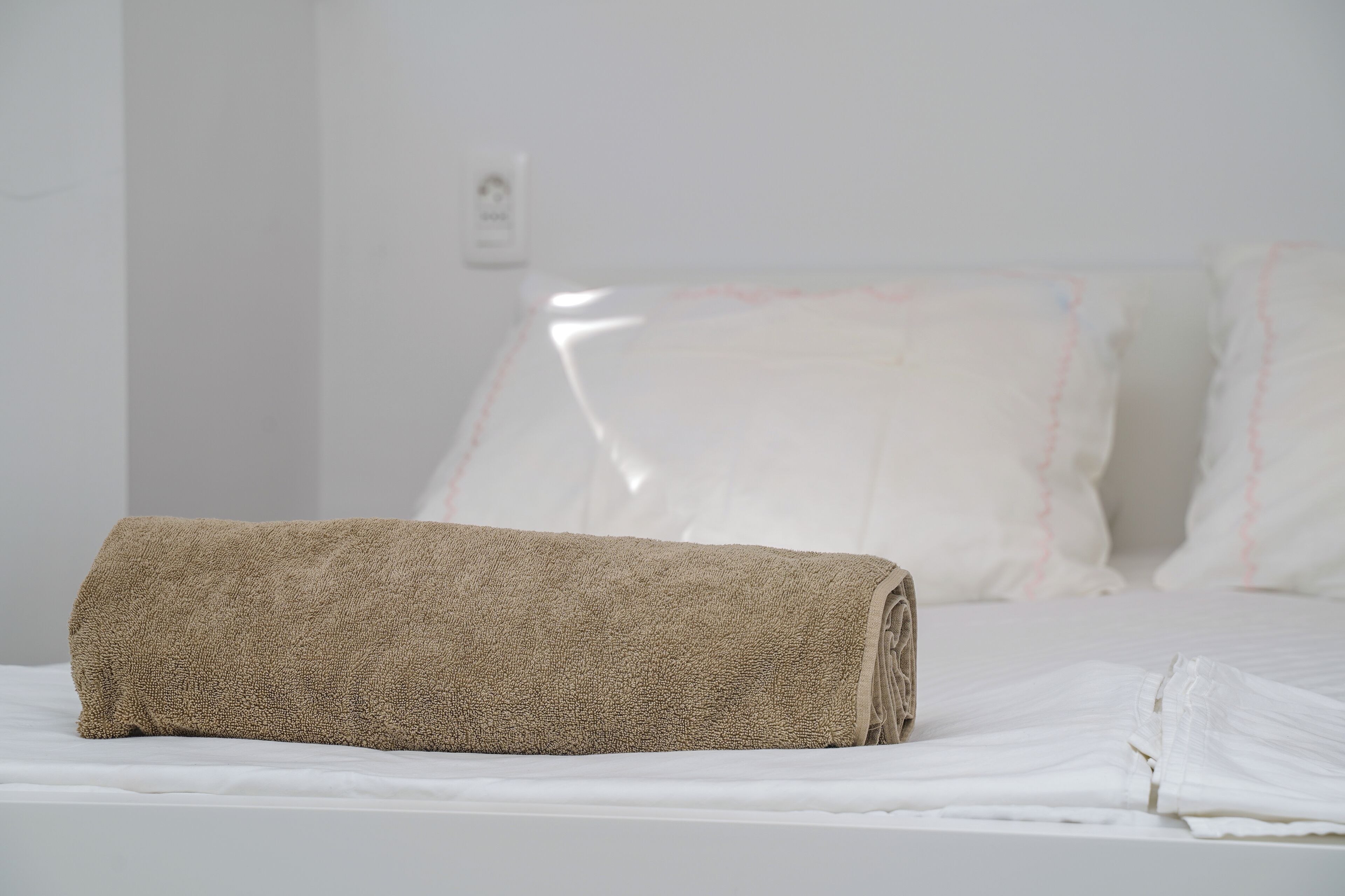 A neatly made bed with white linens and a rolled beige towel on top.
