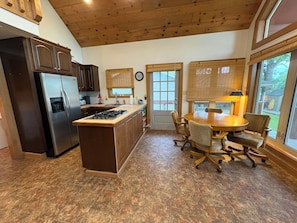 Una cocina con una isla central, armarios de madera, un refrigerador, una mesa de comedor con sillas y ventanas grandes.