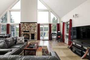 A spacious living room with a stone fireplace, a large TV, and a balcony with red curtains. - Idabel Lake escape , nestled in the mountains near big white (Kootenay Boundary E / West Bo*)