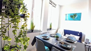 Un comedor con una mesa dispuesta para dos personas, plantas en macetas y un sofá azul al fondo.