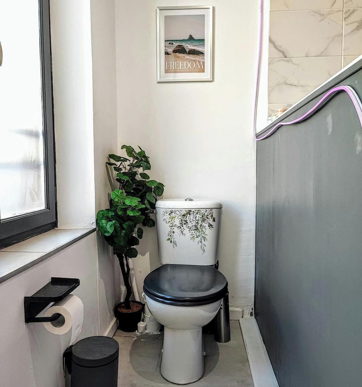 A small bathroom with a toilet, a toilet paper holder, a trash bin, a potted plant, and a framed picture on the wall.