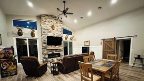 A spacious living room with a stone fireplace, leather sofas, a wooden dining table, and a TV mounted on the wall. - Finger Lake Retreat (Prattsburgh)