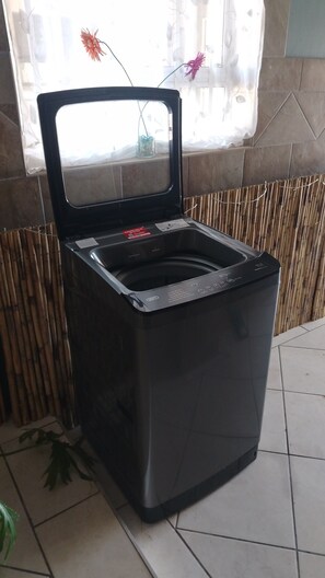 Une machine à laver compacte, noire, à chargement frontal, dotée d’une porte transparente, posée sur un sol carrelé, adossée à un mur en bambou.