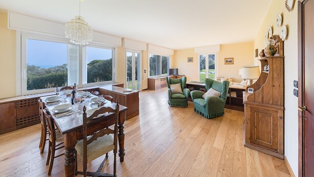 A spacious dining area with a wooden table set for six, large windows offering a view of the outdoors, and a cozy seating area with green upholstered chairs. - Villa Elisa with pool (Padenghe sul Garda)
