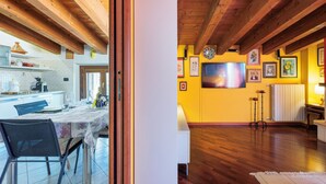 Una stanza con travi a vista in legno, un'area pranzo con tavolo e sedie, e una cucina visibile attraverso una porta aperta.