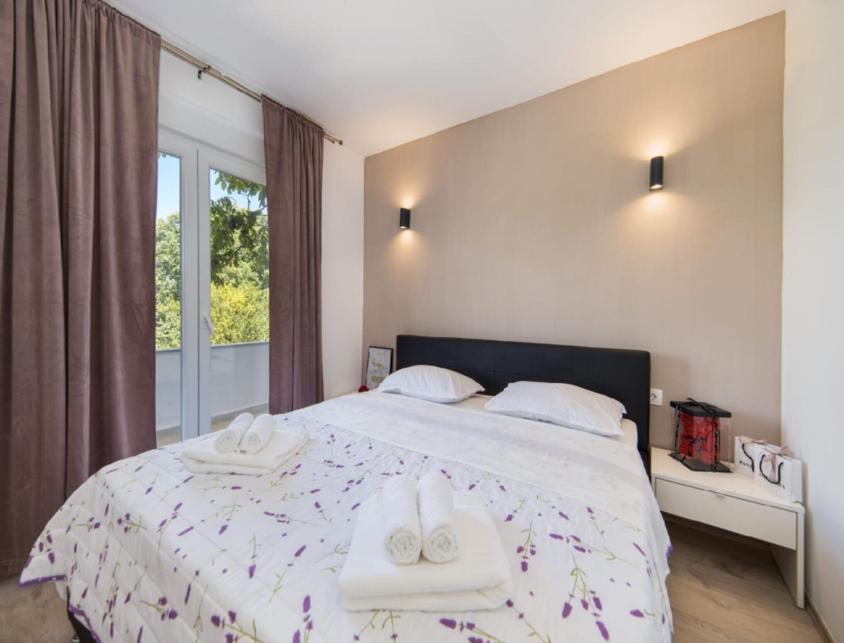 A bedroom with a bed, two bedside tables, a window with curtains, and a view of greenery outside.