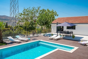 En swimmingpool med liggestole, et moderne hus med et flisebelagt tag og et bjergrigt landskab i baggrunden.